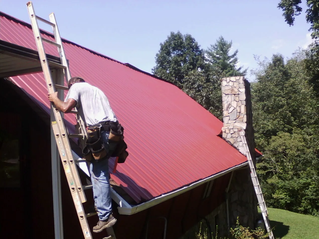 Roofer on a ladder beside a red metal roof and stone chimney — Soffit & Fascia Repair by Roofing Brothers in Balm
